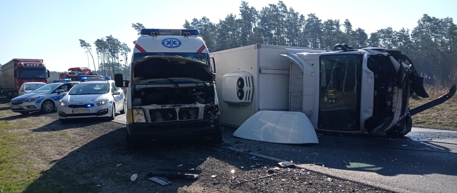 Na zdjęciu widać leżącego na boku uszkodzonego busa oraz stojącą obok uszkodzoną karetkę. W tle dwa radiowozy policji.