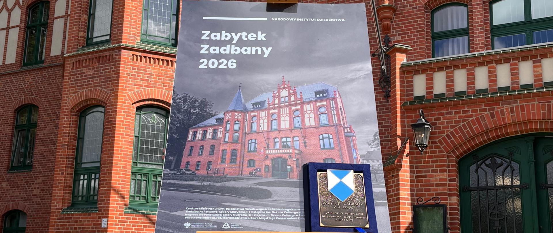 Tablica informacyjna konkursu " Zabytek Zadbany 2026" i oprawiony dyplom na sztaludze przed ceglanym budynkiem Państwowej Szkoły Muzycznej I i II st. im. Oskara Kolberga w Szczecinku. 