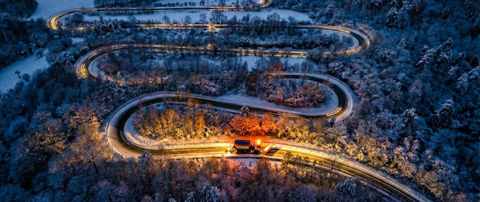 Nocne, zimowe ujęcie z drona przedstawiające krętą, oświetloną serpentynę drogową biegnącą przez las pokryty śniegiem; światła pojazdów tworzą jasne smugi na zakrętach.