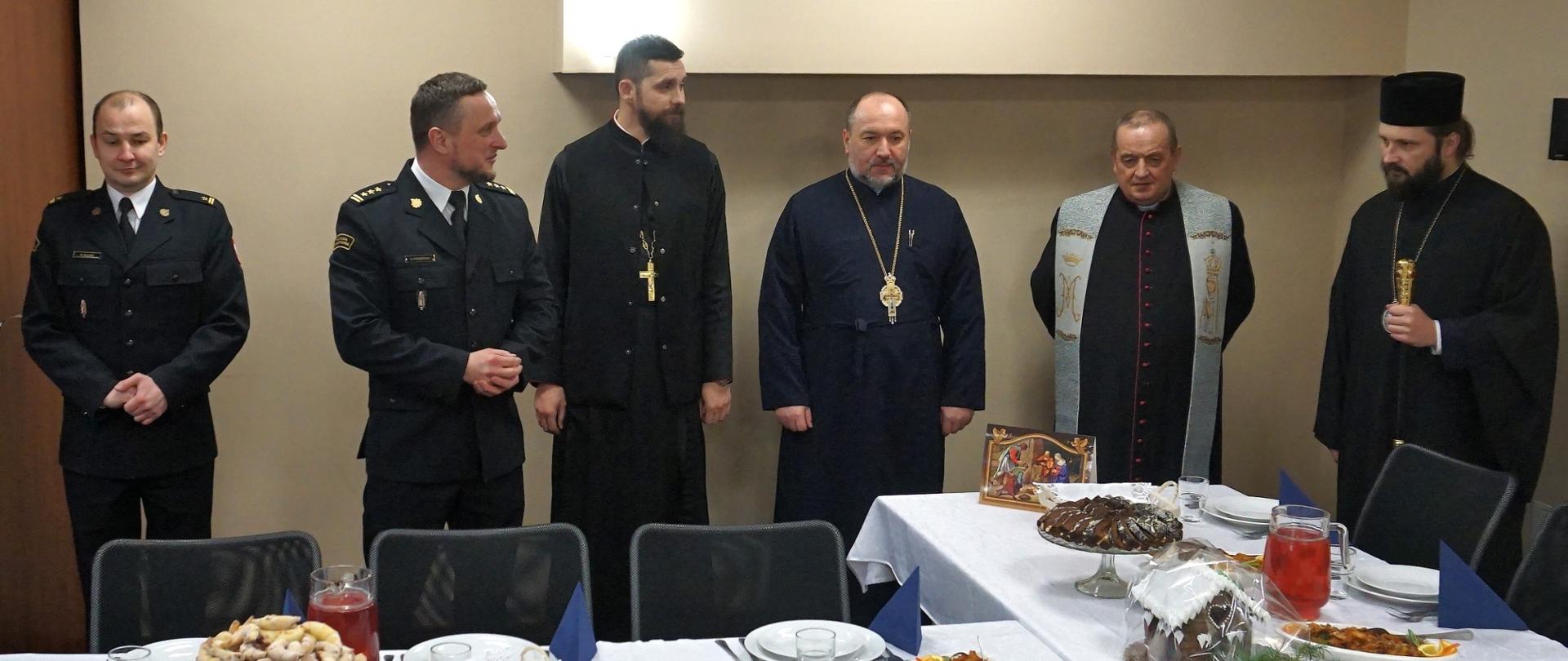 Przy wigilijnym stole stoją osoby duchowne w sutannach i dwóch strażaków w mundurach.