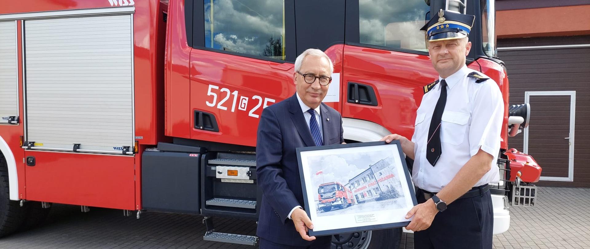 Zdjęcie przedstawia Pana Kazimierza Smolińskiego – Posła na Sejm RP wraz z Komendantem powiatowym na tle samochodu pożarniczego z pamiątkowa grafiką.
