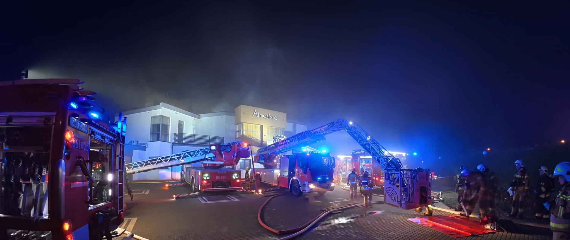 „Akcja gaśnicza prowadzona nocą przez straż pożarną przed budynkiem Atmosfera – widać kilka wozów strażackich z rozłożonymi drabinami, strażaków w pełnym umundurowaniu oraz unoszący się dym nad budynkiem.”