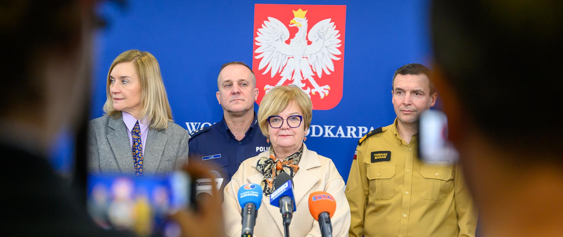 Konferencja prasowa w Podkarpackim Urzędzie Wojewódzkim. W centrum stoi wojewoda podkarpacki Teresa Kubas-Hul. Obok niej przedstawiciele służb mundurowych, w tym nadbryg. Tomasz Baran, podkarpacki komendant wojewódzki PSP, w piaskowym mundurze służbowym. W tle widoczne godło Polski na niebieskim ścianko-panelu z napisem „WOJEWODA PODKARPACKI”. Na pierwszym planie sylwetki dziennikarzy z mikrofonami.