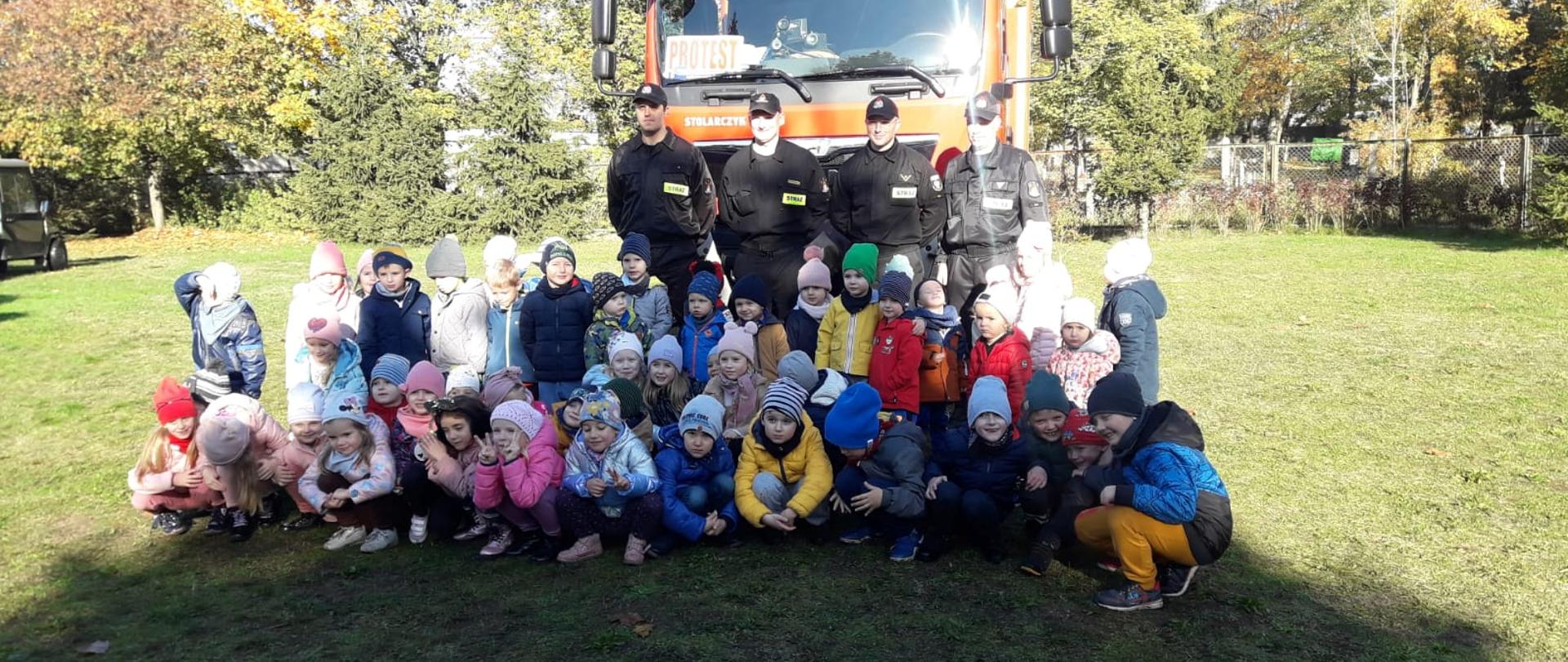 Wspólne zdjęcie przedszkolaków ze strażakami, w tle samochód pożarniczy.