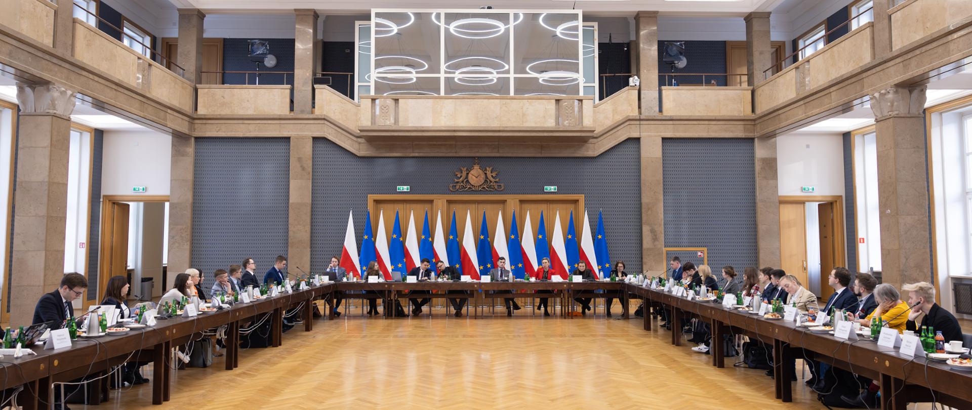 Szeroki kadr. Uczestnicy posiedzenia Rady obradują w sali konferencyjnej przy stole ustawionym w literę U. W tle widoczne flagi Polski i Unii Europejskiej, po bokach widać sali kolumny i okna.