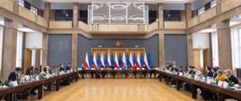 Szeroki kadr. Uczestnicy posiedzenia Rady obradują w sali konferencyjnej przy stole ustawionym w literę U. W tle widoczne flagi Polski i Unii Europejskiej, po bokach widać sali kolumny i okna.