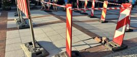 Na zdjęciu widoczne wybrzuszenie terenu w postaci uniesionej kostki otoczone pachołkami z taśmą ostrzegawczą. W tle widoczni przechodnie. Kostka koloru szarego z ceglanym wzorem.