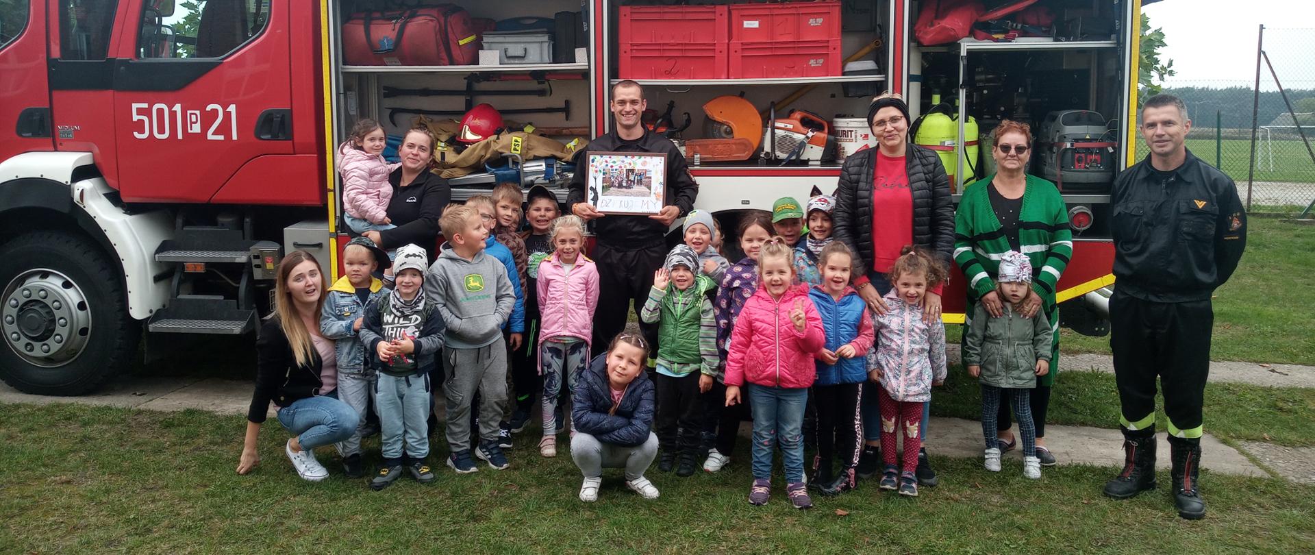 Na zdjęciu dzieci z wychowawcą i strażacy a w tle samochód pożarniczy.