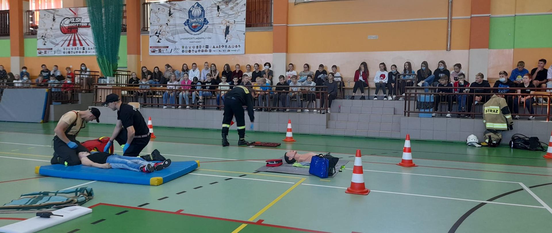 Zdjęcie przedstawia zajęcia w ramach prewencji społecznej światowego dnia pierwszej pomocy w Szkole Podstawowej im. Mariana Falskiego w Kraszewicach. Na zdjęciu widzimy strażaków, którzy przeprowadzają pokaz kwalifikowanej pierwszej pomocy na przykładzie jednego z uczniów szkoły podstawowej. Można też zauważyć dużo sprzętu, który używany jest przez straż pożarną na zdarzeniach. Cały pokaz obserwują na drugim planie uczniowie szkoły. Zdjęcie wykonane w porze dziennej na hali sportowej.