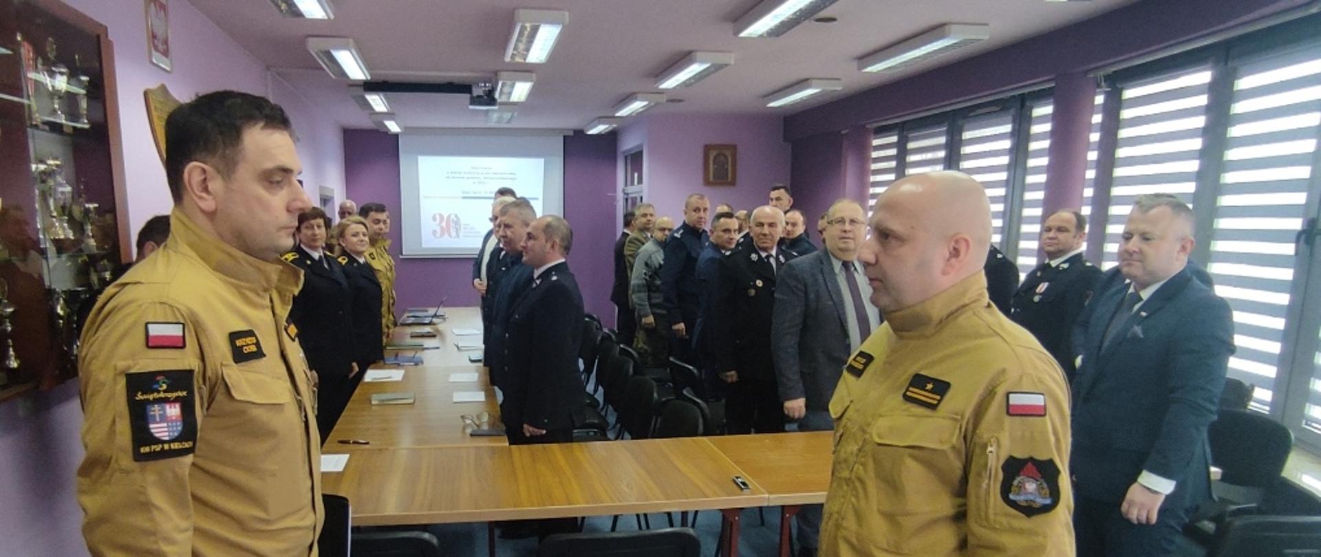 Sala narad KP PSP, przy stołach ustawionych w podkowę stoją przedstawiciele jednostek OSP, służb współdziałających oraz samorządowcy, dowódca JRG składa meldunek Świętokrzyskiemu Komendantowi Wojewódzkiemu PSP