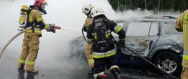 Zdjęcie przedstawia pożar samochodu osobowego na autostradzie A2. Z samochodu wydobywa się gęsty dym. Strażacy podają wodę oraz pianę gaśniczą na palący się pojazd. 