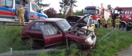 Fotografia przedstawia uszkodzony pojazd osobowy w przydrożnym rowie. W tle widać ratowników PSP i ich pojazdy oraz ZRM. 