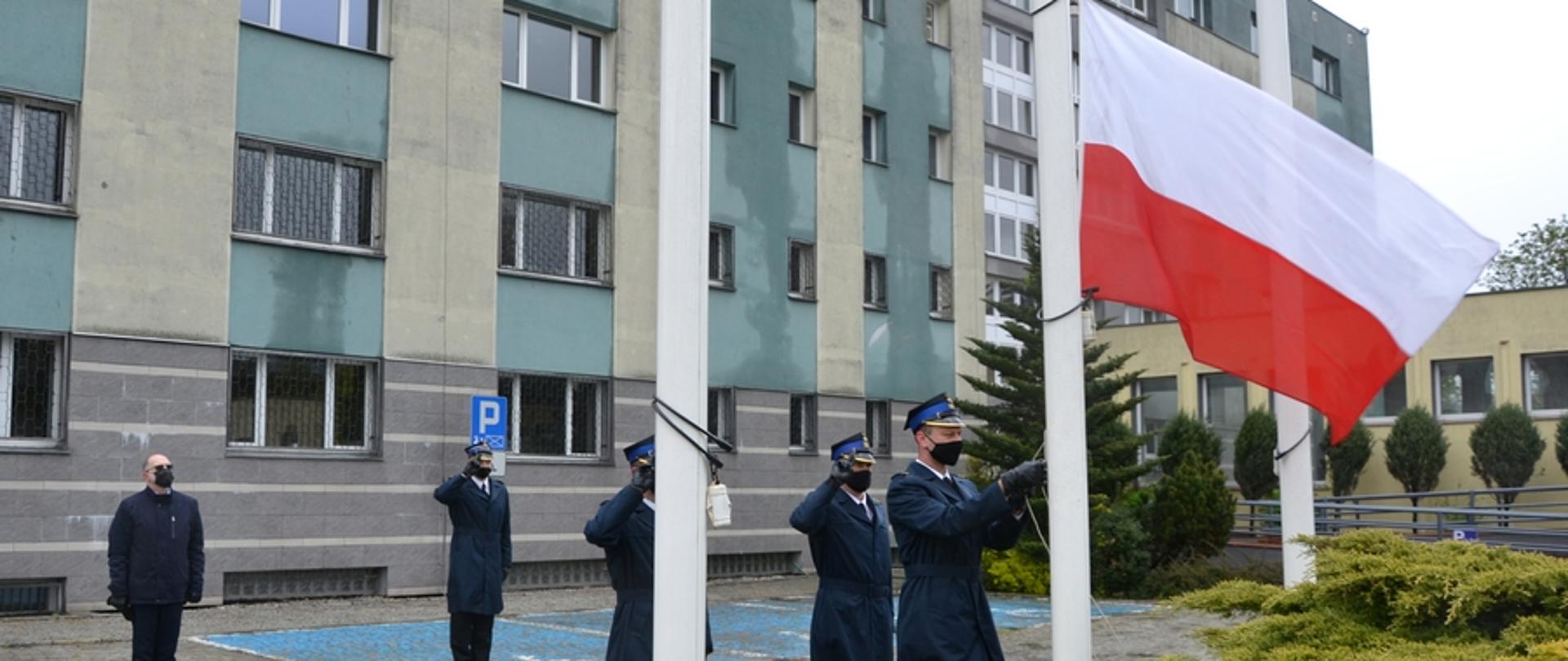 Na zdjęciu widać strażaków w mundurach wyjściowych, którzy w uroczysty sposób podnoszą na maszt flagę państwową z okazji Dnia Flagi Rzeczypospolitej Polskiej. W tle budynek starostwa powiatowego