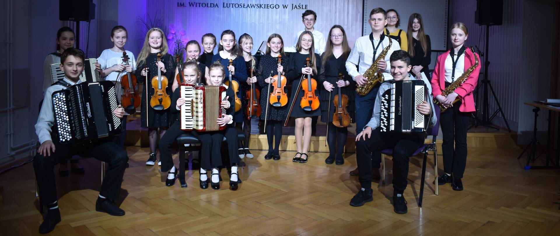Na zdjęciu widnieje grupa uczniów, ustawionych na scenie auli PSM I stopnia w Jaśle. Uczniowie trzymają w rękach skrzypce, saksofony i akordeony. Przeważająca kolorystyka zdjęcia: brązowo-czarno-biała.