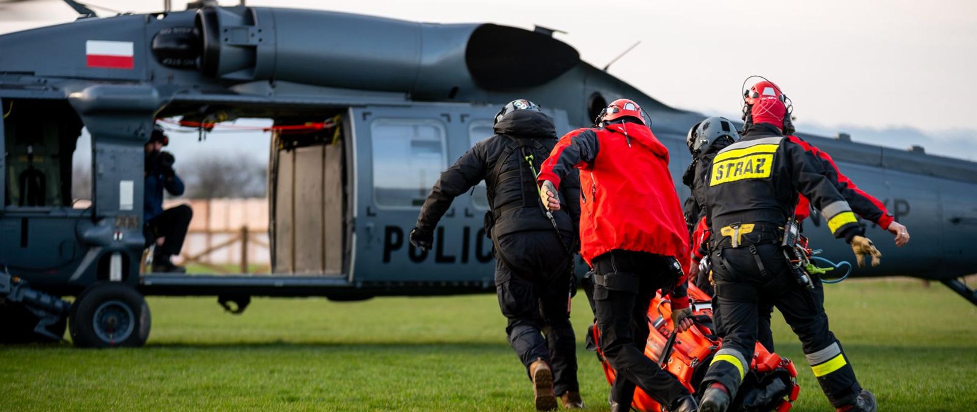 Strażacy ratownicy niosą zbiornik podwieszany typu bumbi bucket w kierunku stojącego na lądowisku śmigłowca S70i Black Hawk Policji