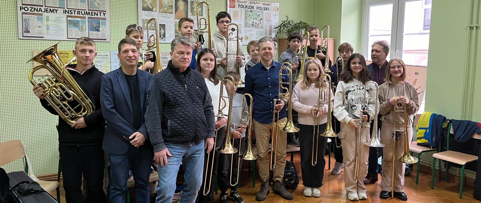 Na zdjęciu grupa osób, którzy trzymają puzony. Na podłodze drewniany parkiet. a ściany w kolorze oliwkowo zielonym.