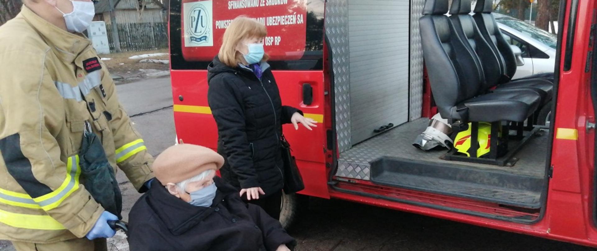 Zdjęcie przedstawia pojazd typu bus należący do jednostki Ochotniczej Straży Pożarnej w Jedlni Letnisko. Pojazd w kolorze czerwonym z białymi elementami zabudowy. Tylne drzwi pojazdu są otwarte. Przed pojazdem stoi strażak. Strażak ubrany w ubranie specjalne w kolorze żółtym. Przed nim znajduje się wózek inwalidzki, na którym siedzi starsza osoba. Pomiędzy nimi a samochodem stoi jeszcze jedna kobieta, opiekunka starszej osoby siedzącej na wózku. Za pojazdem widać drzewa. 
