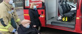 Zdjęcie przedstawia pojazd typu bus należący do jednostki Ochotniczej Straży Pożarnej w Jedlni Letnisko. Pojazd w kolorze czerwonym z białymi elementami zabudowy. Tylne drzwi pojazdu są otwarte. Przed pojazdem stoi strażak. Strażak ubrany w ubranie specjalne w kolorze żółtym. Przed nim znajduje się wózek inwalidzki, na którym siedzi starsza osoba. Pomiędzy nimi a samochodem stoi jeszcze jedna kobieta, opiekunka starszej osoby siedzącej na wózku. Za pojazdem widać drzewa. 
