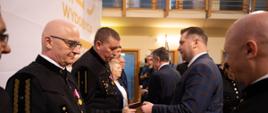 Minister Czarnek stoi przed szeregiem mężczyzn w czarnych galowych mundurach, podaje jednemu z nich małą książeczkę.