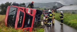 Przewrócony samochód ciężarowy na poboczu a obok niego strażacy. Jeden strażak na samochodzie ciężarowym.