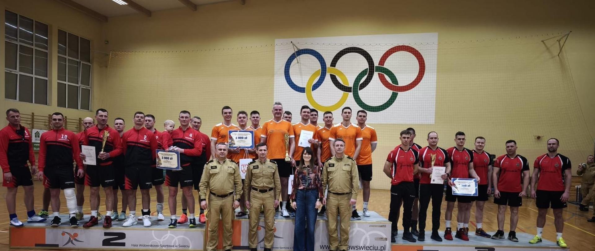 Strażacy z drużyn PSP stoją na podium w hali sportowej podczas ceremonii zakończenia Kujawsko-Pomorskich Mistrzostw Strażaków PSP w Piłce Siatkowej. Na środku podium znajduje się zwycięska drużyna KP PSP Radziejów.