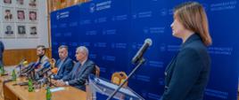 Minister Wieczorek i dwóch mężczyzn w garniturach siedzą za drewnianym stołem, za nimi niebieska ścianka z napisem Uniwersytet Szczeciński, z boku przy mikrofonie na stojaku stoi kobieta w granatowej marynarce.