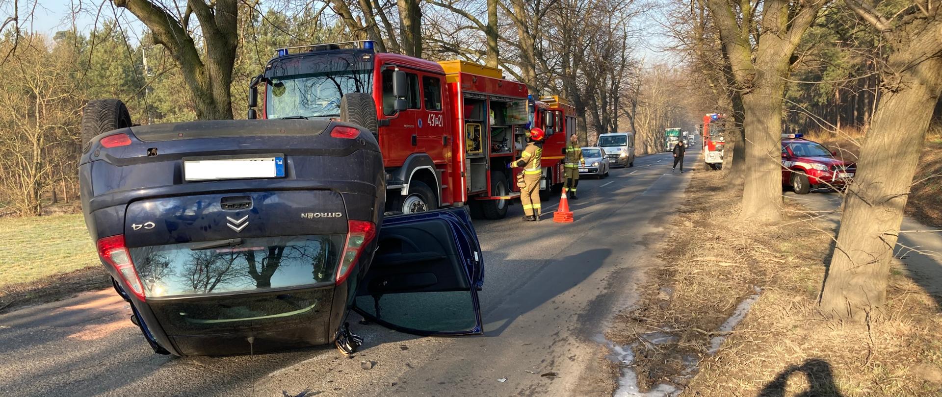 Na pierwszym planie, na dachu leży samochód osobowy. W tle stoją pojazdy pożarnicze.