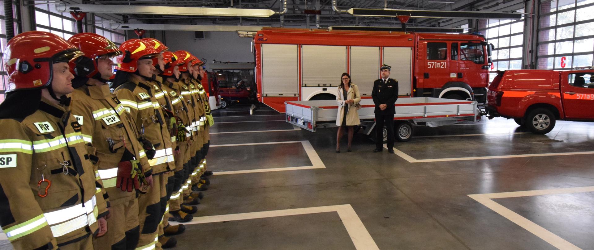 Kobieta w ubraniu cywilnym i mężczyzna w ubraniu strażackim stoją na garażu przed przyczepką zaczepioną do samochodu terenowego. Z prawej szereg strażaków w ubraniach specjalnych.