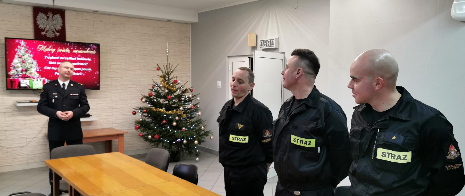 Mężczyźni w ubraniach strażackich koszarowych stojący przy stole na końcu którego stoi przemawiający komendant powiatowy. W oddali przystrojona choinka.