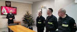 Mężczyźni w ubraniach strażackich koszarowych stojący przy stole na końcu którego stoi przemawiający komendant powiatowy. W oddali przystrojona choinka.