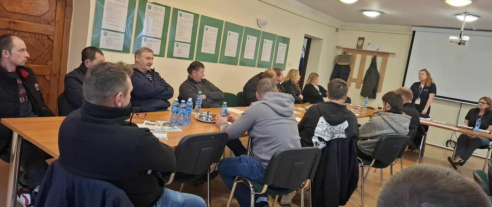 Uczestnicy spotkania siedzący przy długiej ławce, kobieta stojąca naprzeciw uczestników.