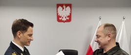 Komendant Powiatowy PSP w umundurowaniu wyjściowym wręcza pamiątkowe życzenia nagrodzonemu funkcjonariuszowi. w tle szara ściana, na której wisi godło RP. Panowie podają sobie ręce.