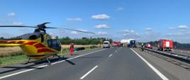 Na autostradzie 4 po lewej na pierwszym planie stojący śmigłowiec LPR. Pilot udaje się w kierunku zdarzenia. Na dalszym planie – po lewej w rowie widać dwa pojazdu uczestniczące w zdarzeniu. W środku widać karetkę oraz samochody straży pożarnej. Przy barierkach oddzielających pasy ruchu w oddali widać samochód dostawczy w którego tył uderzył ciągnik siodłowy – cementowóz stojący za samochodem dostawczym. Na przeciwległym pasie kolejne dwa pojazdy straży pożarnej- operacyjny pickup i gaśniczy. W tle pobliski las i niebo z niewielkim zachmurzeniem.
