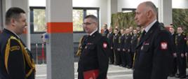 Komendanci zdający i przejmujący obowiązki stoją na przeciw Komendanta Wojewódzkiego, trzymają w rękach teczki. Za nimi w tle strażacy w mundurach.