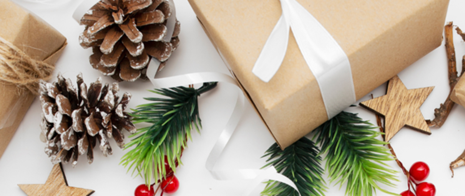 Na zdjęciu widać elegancką, świąteczną grafikę.
U góry kompozycji znajdują się zapakowane prezenty w brązowy papier, przewiązane białymi wstążkami. Towarzyszą im naturalne dekoracje: szyszki, gałązki świerku, czerwone jagody oraz drewniane gwiazdki.