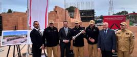 4 strażaków PSP oraz 3 mężczyzn w garniturach stoi przed murami budowanej strażnicy, jeden funkcjonariusz trzyma w ręku tubę z aktem erekcyjnym, po lewej stronie na sztaludze ustawiony plakat z wizualizacją budowanego obiektu
