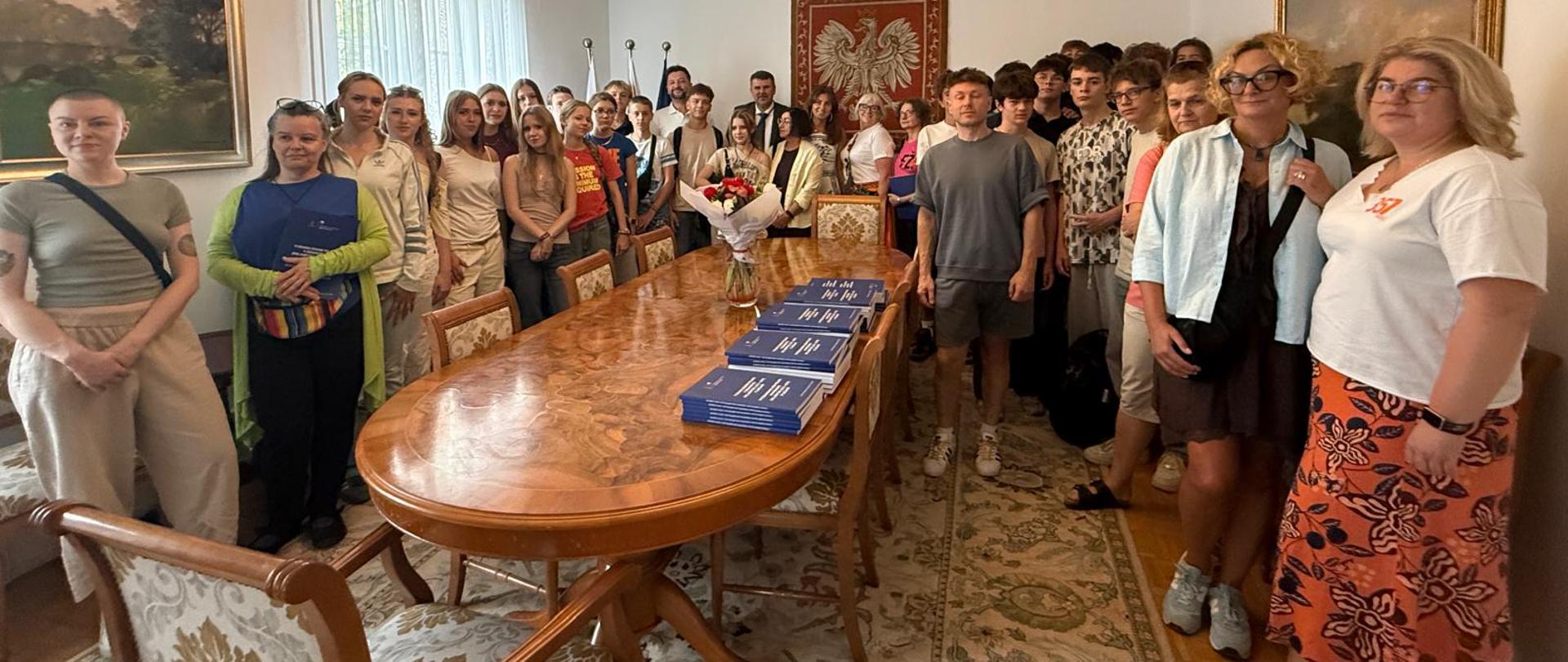 Večja skupina mladih in profesorjev pozira za skupinsko fotografijo v prostorih Veleposlaništva Republike Poljske v Ljubljani. Udeleženci stojijo okoli dolge mize, na steni za njimi pa je poljski državni grb.