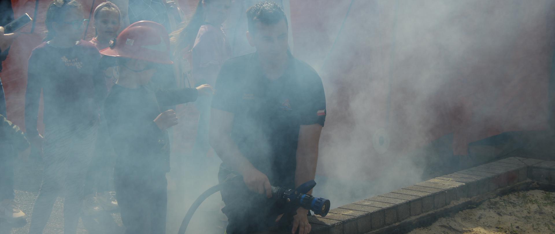 Dzień otwarty w rawickiej komendzie. Na "stanowisku gaśniczym" znajduje sie strażak trzymający w ręce prądownicę, oraz jego pomocnik w czerwonym hełmie. Za nimi obserwatorzy. Teren jest zadymiony.