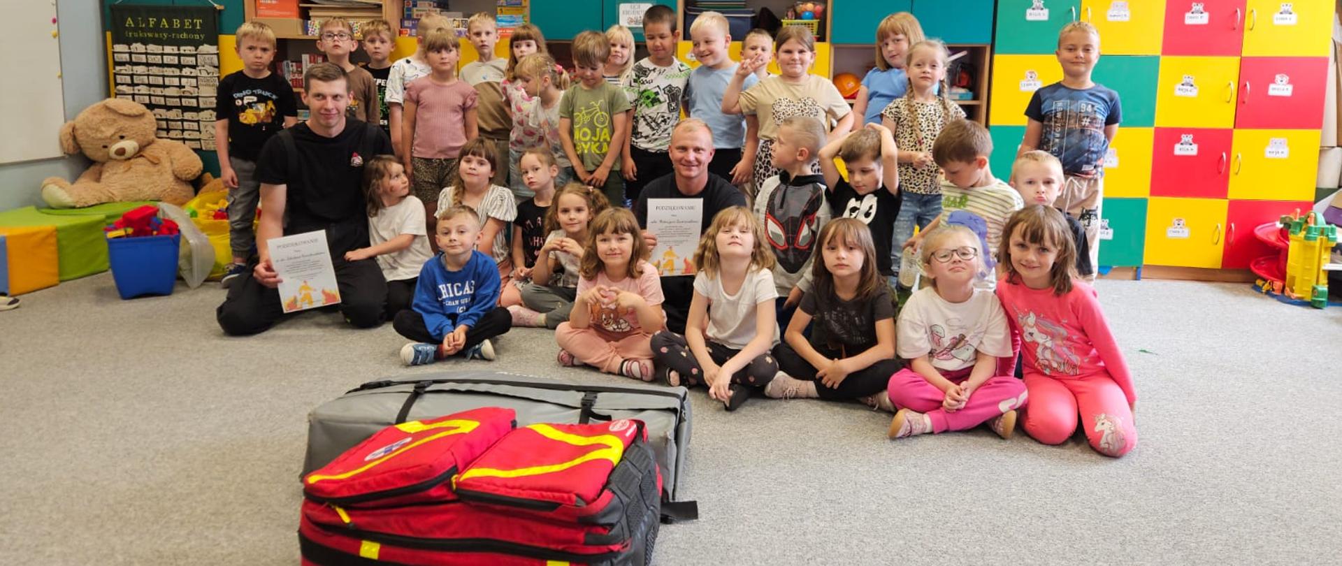 Zdjęcie przedstawia dzieci klas 0 Szkoły Podstawowej nr 2 w Gołdapi pozujące wspólnie z dwoma strażakami po spotkaniu edukacyjnym, którzy są zlokalizowani w jednej z klas