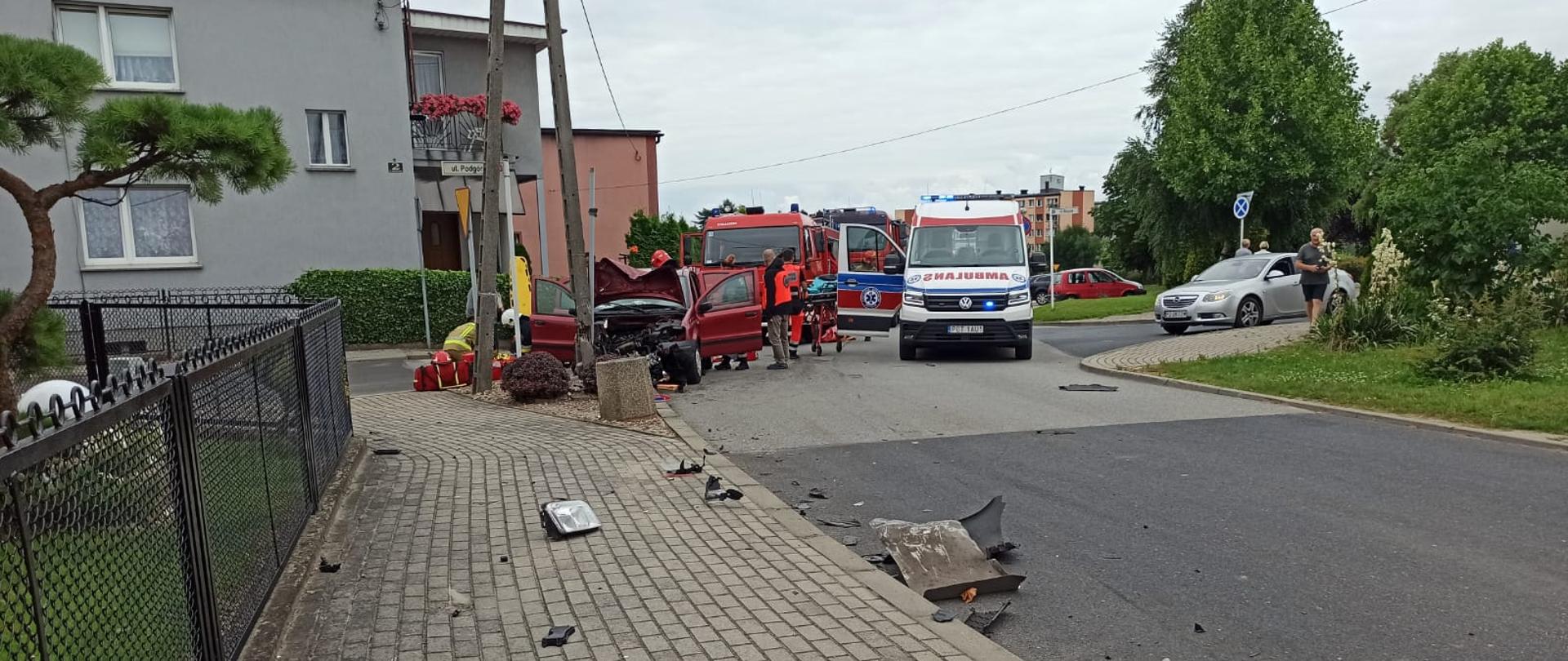 Na pierwszym planie rozrzucone elementy z rozbitych pojazdów. na drugim widać rozbity pojazd, który uderzył w słup energetyczny. Obok karetka, radiowóz policyjny nieoznakowany, W tle samochód pożarniczy, po lewej jego stronie budynku mieszkalne.