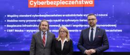 Na tle wielkiego niebieskiego ekranu z napisem Nowelizacja ustawy o Krajowym Systemie Cyberbezpieczeństwa stoi mężczyzna w szarym garniturze, kobieta w czarnej marynarce i minister Kulasek w ciemnoszarym garniturze.