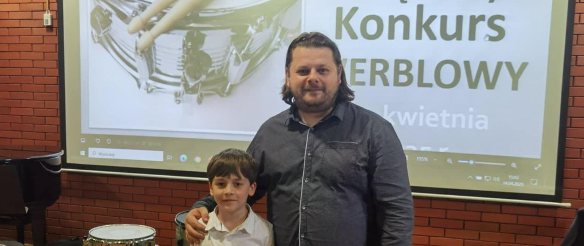 Chłopiec z dyplomem pozuje razem z dorosłym mężczyzną – prawdopodobnie nauczycielem lub opiekunem – na tle ekranu z banerem konkursu. Oboje uśmiechnięci, stoją obok siebie w sali koncertowej.