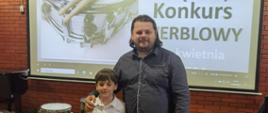 Chłopiec z dyplomem pozuje razem z dorosłym mężczyzną – prawdopodobnie nauczycielem lub opiekunem – na tle ekranu z banerem konkursu. Oboje uśmiechnięci, stoją obok siebie w sali koncertowej.