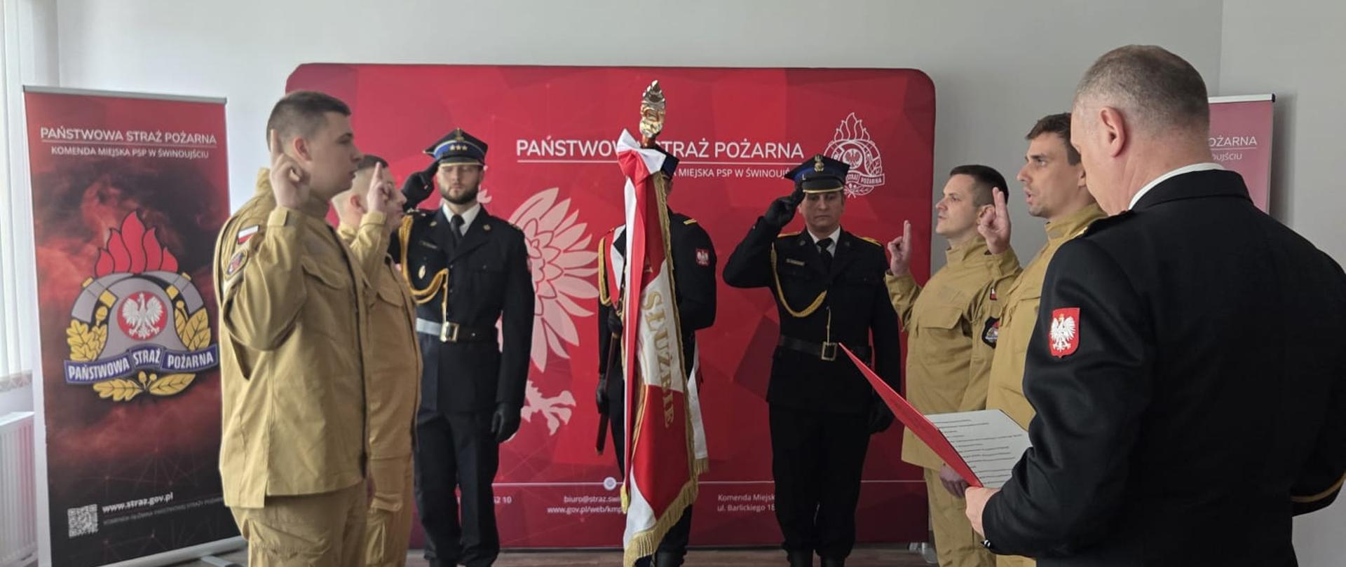 zdjęcie przedstawia strażaków przystających do ślubowania. W tle widoczny poczet sztandarowy w umundurowaniu wyjściowym koloru granatowego. Sztandarowy pochyla sztandar w przód, a ślubujący w umundurowaniu służbowym koloru piaskowego z jedną ręką uniesioną w górę powtarzają rotę ślubowania. Po prawej stronie zdjęcia widoczny przyjmujący ślubowanie.