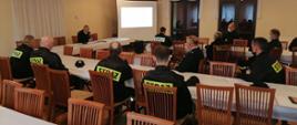 Na zdjęciu sala w której siedzą przy stołach w odstępach strażacy w umundurowaniu koszarowym; strażacy widziani z boku. Jeden strażak widziany z przodu siedzi przy stole prezydialnym, trzyma mikrofon. W tle ekran do wyświetlania.