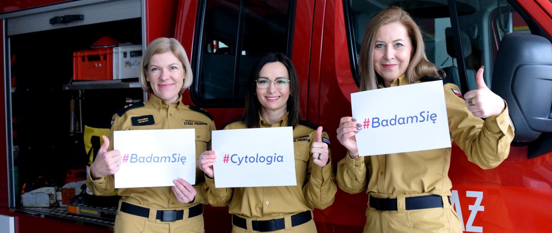 Zdjęcie zrobione w garażu jednostki ratowniczo-gaśniczej. Na pierwszym planie stoją trzy funkcjonariuszki Państwowej Straży Pożarnej w piaskowych mundurach. W jednej ręce trzymają kartki z nadrukami: #BadamSię oraz #Cytologia. Druga ręka jest z uniesionym kciukiem. Za nimi ustawiony jest samochód strażacki.