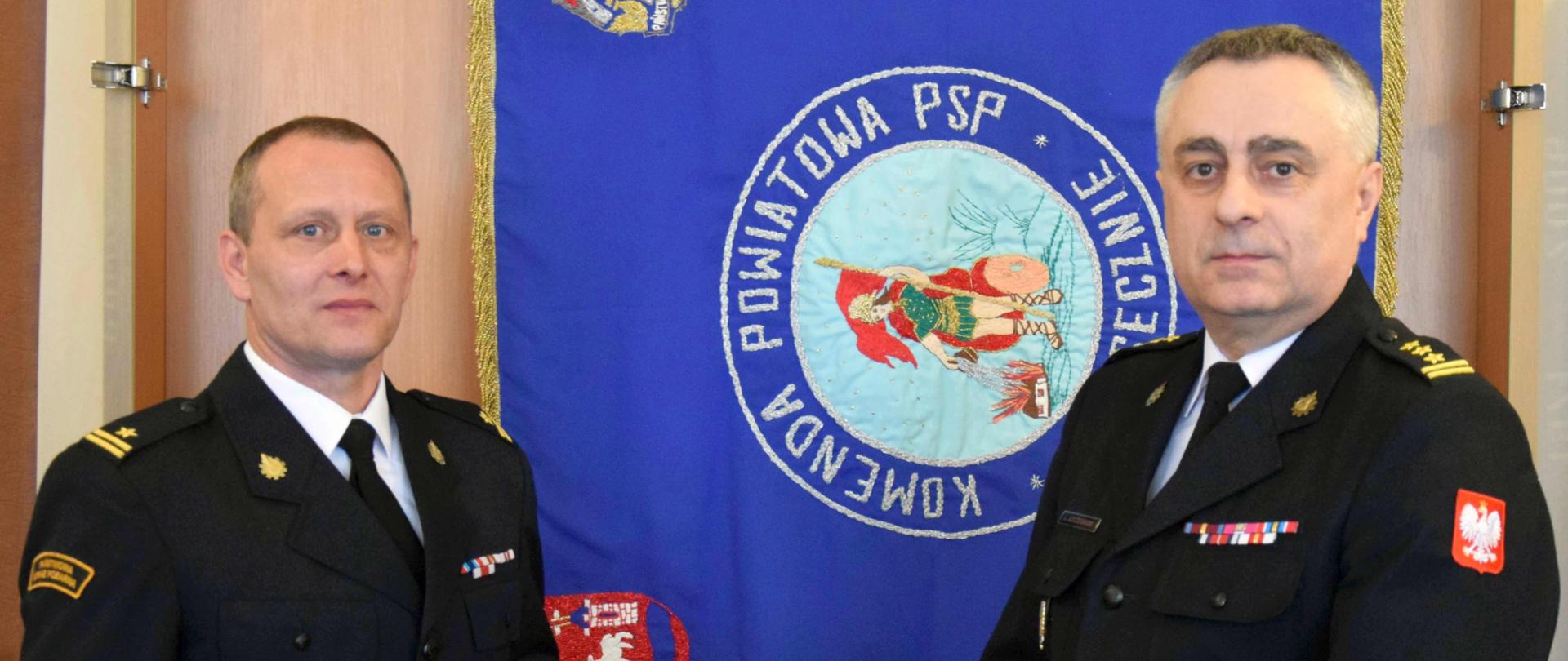 P.o. Zastępcy Komendanta Powiatowego PSP w Piasecznie trzyma w ręku dokument powierzający pełnienie obowiązków. Obok stoi komendant Powiatowy PSP. W tle sztandar Komendy Powiatowej PSP w Piasecznie 