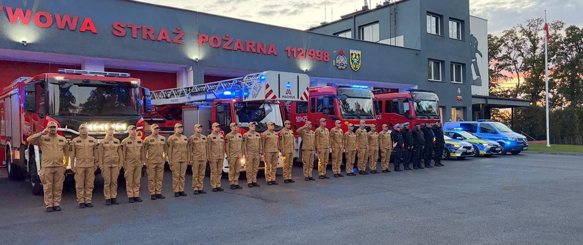 Osoby w mundurach strażackich oraz policjanci stoją w szeregu przed budynkiem Państwowej Straży Pożarnej. Za nimi zaparkowane są czerwone wozy strażackie z drabiną oraz niebiesko-srebrne radiowozy policyjne z włączonymi sygnałami świetlnymi.