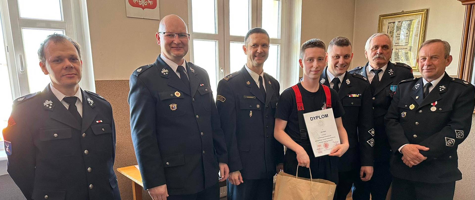 Zdjęcie przedstawia strażaków, dzieci i młodzież podczas turnieju wiedzy z zakresu ochrony przeciwpożarowej.
W tle pomieszczenie.
