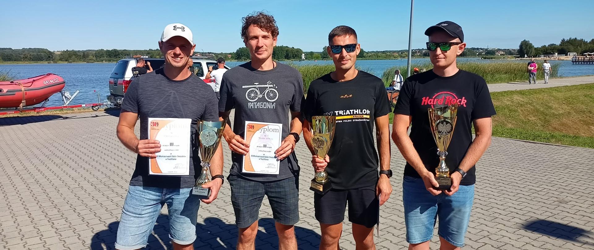 Reprezentacja KM PSP Opole na 3. Mistrzostwach Polski Strażaków PSP w ramach IX edycji Triathlonu Kraśnik – Przegrywają Jedynie Nieobecni. W strażacy w ubraniach cywilnych pozują do zdjęcia trzymając trzy puchary i dwa dyplomy. Funkcjonariusze zdobyli trzecie miejsce w kategorii sztafet PSP. W tle widoczny akwen oraz samochód z pontonem.
